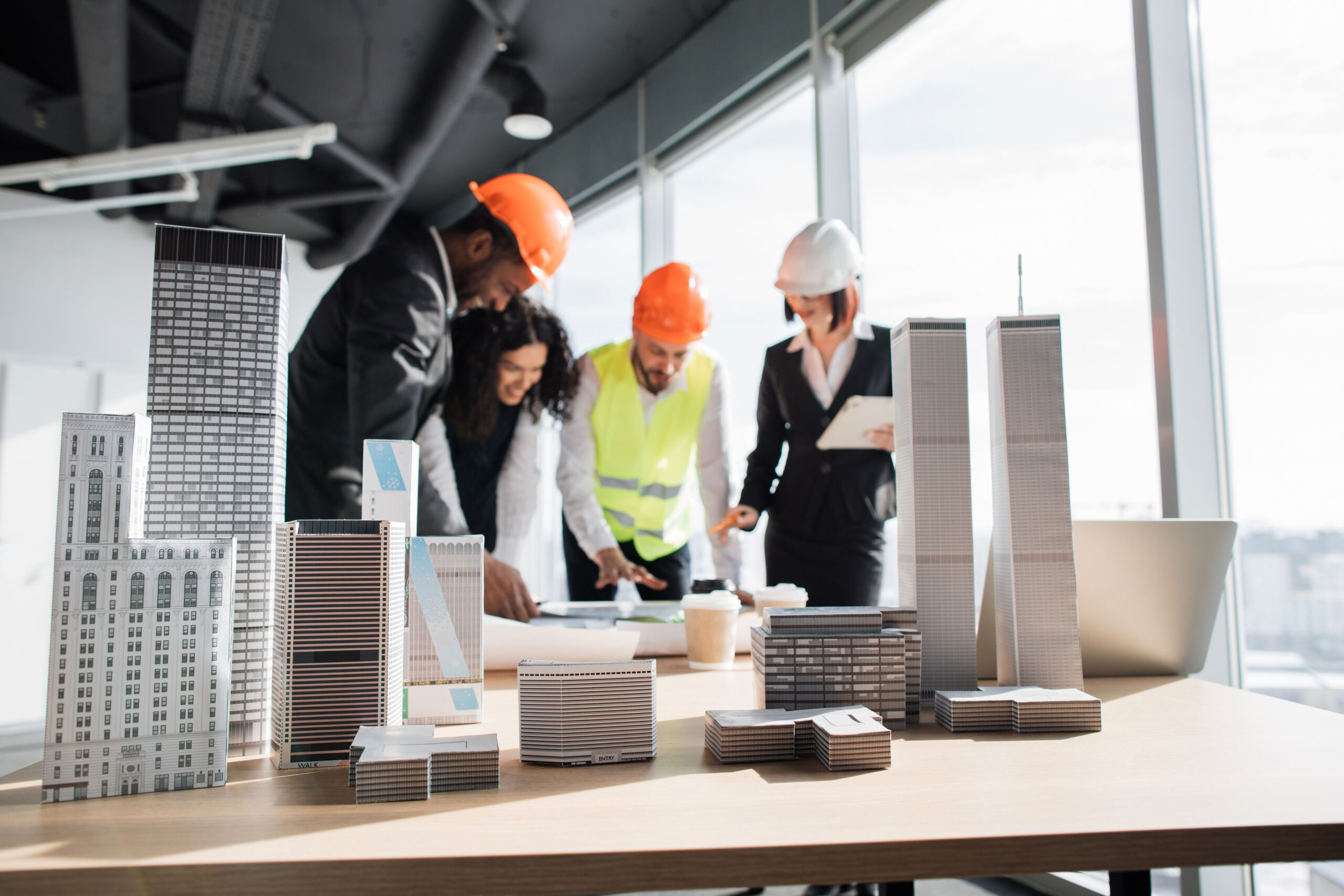 Focus on architecture design of buildings with residential project maquette, group of four multiracial people architects standing near desk with lots of blueprints and at office with panoramic windows